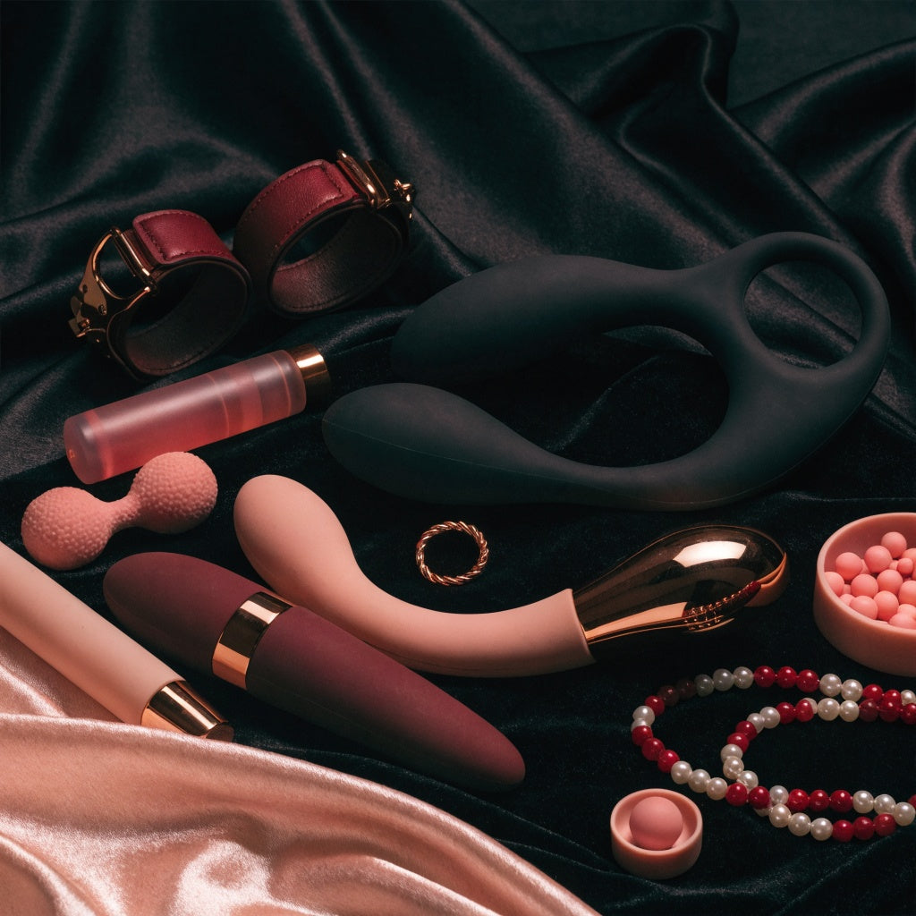 Premium sexual stimulators and intimate wellness accessories in matte black, blush pink and deep burgundy with rose-gold accents, elegantly arranged on luxurious dark satin.