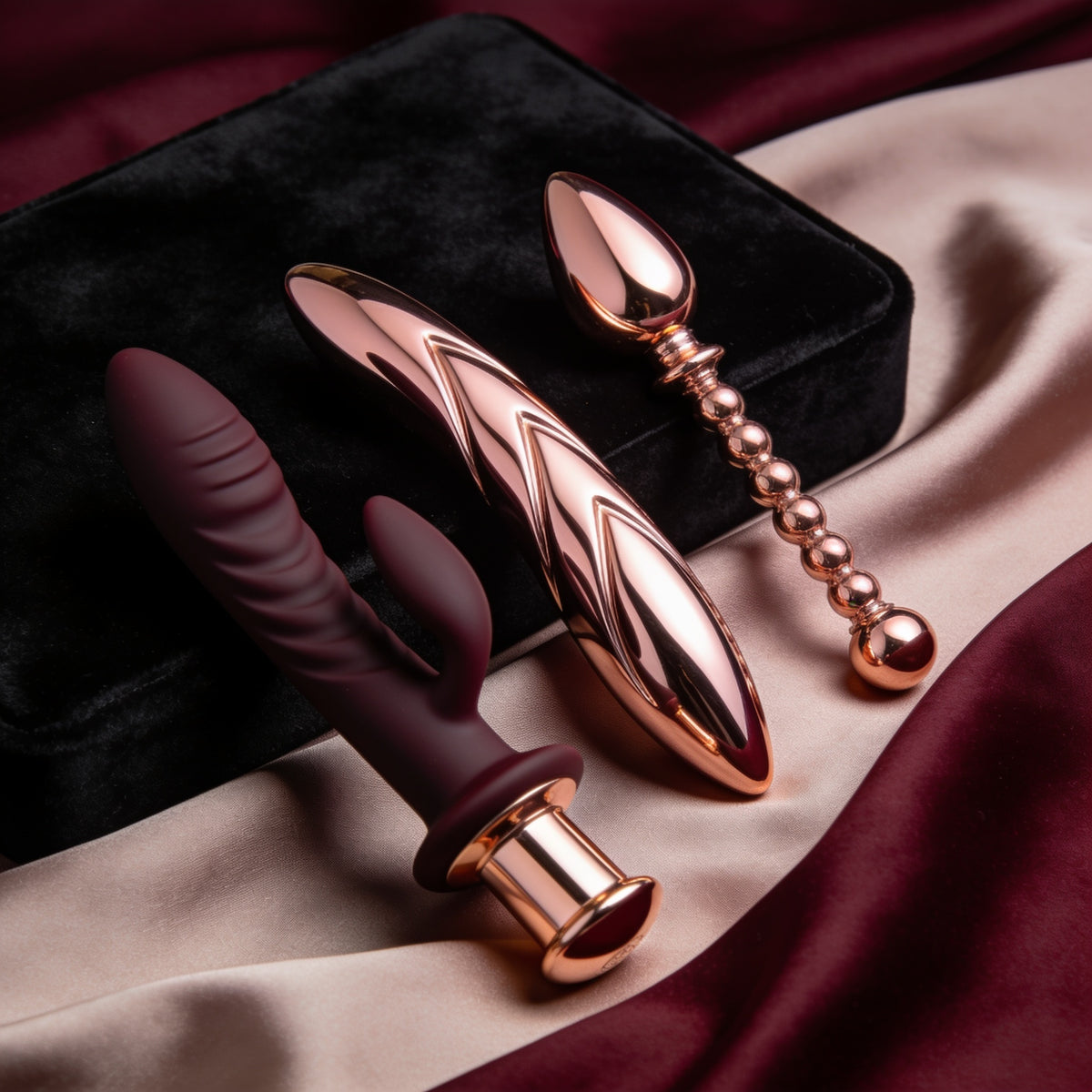 Luxury rose gold sexual wellness toys including a rabbit vibrator, sleek personal massager and beaded pleasure wand displayed on burgundy and blush satin.